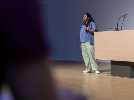 A woman is standing on a stage. She has black hair, a blue blouse, green trousers and white trainers. She holds a microphone in her hand and speaks to the audience.