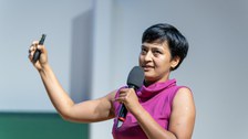 A woman with a pink short-sleeved blouse and short black hair. She holds a microphone in one hand and a presenter in the other.