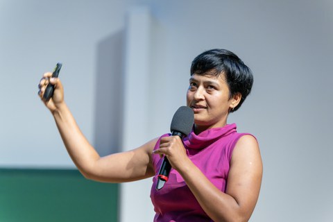 A woman with a pink short-sleeved blouse and short black hair. She holds a microphone in one hand and a presenter in the other.