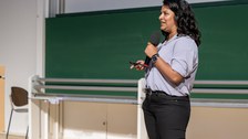 A woman with long black hair, a short-sleeved light blue blouse and black trousers stands on a stage. She holds a microphone in one hand and a presenter in the other.