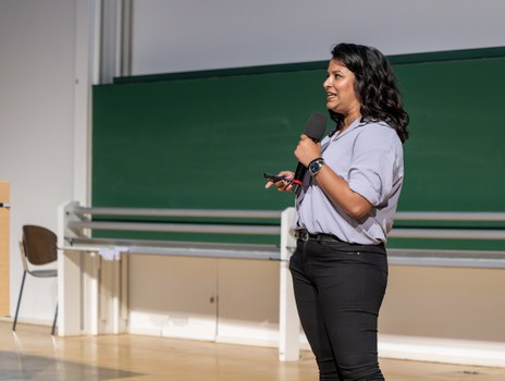 A woman with long black hair, a short-sleeved light blue blouse and black trousers stands on a stage. She holds a microphone in one hand and a presenter in the other.
