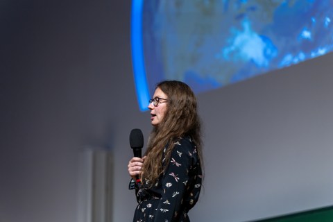 A woman with long light brown hair and glasses speaks into a microphone. She is wearing a black dress that is visible down to her hips. The dress has white appliqués.