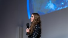 A woman with long light brown hair and glasses speaks into a microphone. She is wearing a black dress that is visible down to her hips. The dress has white appliqués.