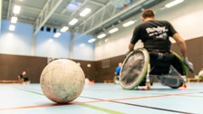 Ein Ball liegt im Vordergrund des Bildes in einer Turnhalle und dahinter sieht man unscharf drei Rollstuhlfahrer beim Sport.
