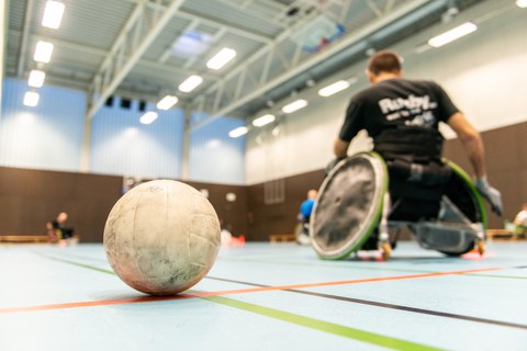Ein Ball liegt im Vordergrund des Bildes in einer Turnhalle und dahinter sieht man unscharf drei Rollstuhlfahrer beim Sport.
