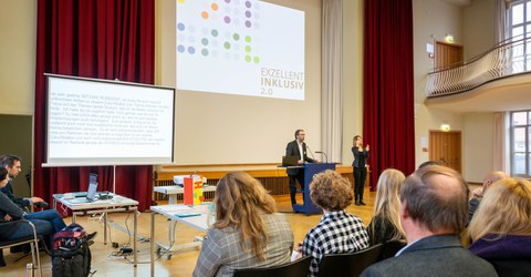Das Foto zeigt einen Vortrag im Dülfersaal der TU Dresden aus der Perspektive des Publikums. Im Hintergrund sind der Referent und eine Gebärdendolmetscherin zu sehen, die vor einer Präsentation mit dem Titel „Exzellent Inklusiv 2.0“ stehen. Links im Bild 