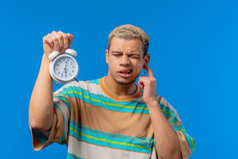 A young man is holding an alarm clock in his right hand whilst covering his ear with his left. He looks confused and is wearing a striped T-shirt. 