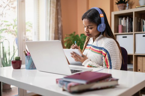 The photo shows a person sitting at a desk. They are  wearing headphones, have a laptop open in front of them, and are holding a notepad in their hand, on which they are writing. There are books lying on the edge of the desk. 