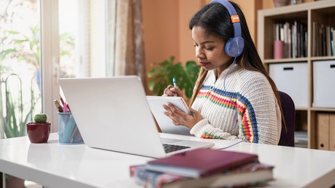 Auf dem Foto ist eine Person an einem Schreibtisch zu sehen. Sie hat Kopfhörer auf, vor ihr steht ein aufgeklappter Laptop und in ihrer Hand ist ein Block auf den sie schreibt. Am Rand des Tisches liegen Bücher. 