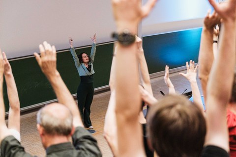 Beim Science Slam 2025 in Dresden steht eine Vortragende vorne im Hörsaal und animiert das Publikum, die Arme zu heben. Viele Zuhörer machen mit, wodurch eine lebendige, interaktive Stimmung entsteht.