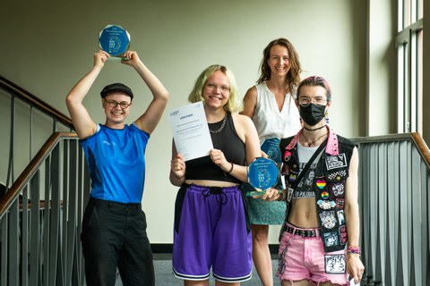 Four people standing on stairs; two holding awards, one holding a certificate.
