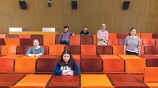 Das Foto zweigt eine Gruppe von 6 Personen, die in einem Vorlesungsaal mit orangenen Sitzgelegenheiten verteilt auf unterschiedliche Reihen sitzen oder stehen. 