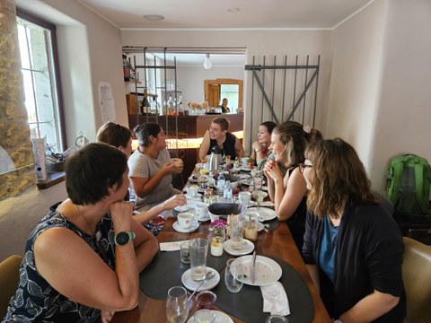Mehrere Frauen sitzen an einer langen Tafel in einem gemütlichen Restaurant und unterhalten sich beim Frühstück. Auf dem Tisch stehen Teller, Tassen, Gläser und verschiedene Speisen.  