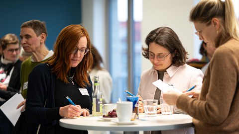 Mehrere Frauen stehen an einem Stehtisch beim Neujahrsempfang des Women@DDc Netzwerks, schreiben Notizen und tauschen sich in entspannter Atmosphäre bei Getränken aus.