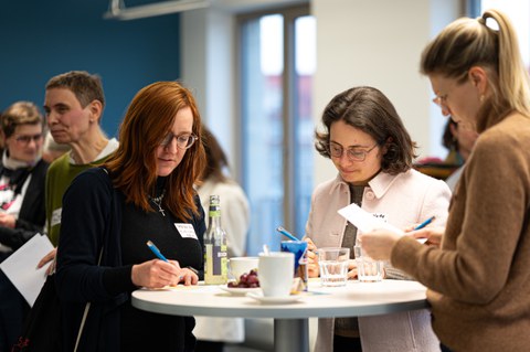 Several women stand at a bar table at the New Year's reception of the Women@DDc network, writing notes and chatting over drinks in a relaxed atmosphere.