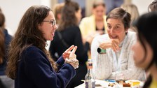 Two women are sitting at a table at the New Year's reception of the Women@DDc network, chatting animatedly and gesturing, while drinks and cake are on the table.
