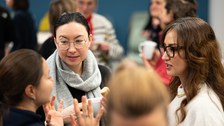 A group of women chat, food and drinks in hand, at the Women@DDc New Year's reception. Other groups of women can be seen in the background. 