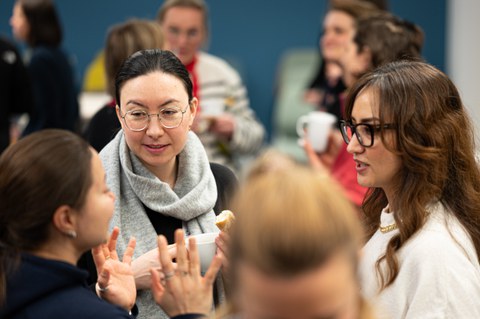 A group of women chat, food and drinks in hand, at the Women@DDc New Year's reception. Other groups of women can be seen in the background. 