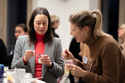 Two women are standing at a bar table, laughing together and eating dessert from a glass with spoons.