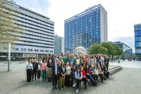 Große Gruppe draußen vor modernen Gebäuden für ein Gruppenfoto versammelt, viele Personen nebeneinander stehend.