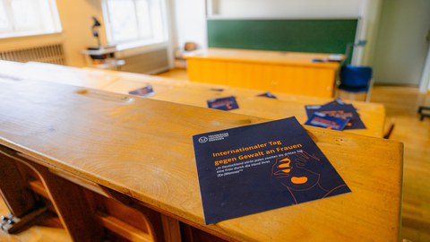 Auf der Sitzbank in eienm Hörsaal liegt das Programmgheft zum Rahmenprogramm des Internationalen Tag gegen Gewalt an Frauen