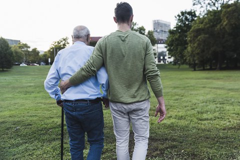 Picture of a grandfather with a cane being supported by his grandson and walking with him across a meadow.