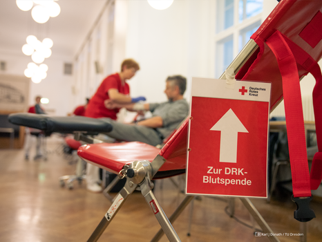 An einem Stuhl hängt ein rotes Schild des deutschen Roten Kreuzes mit einem weißen Pfeil. Darunter ist weißer Schrift zu lesen "Zur Blutspende". Im Hintergrund erkennt man eine Person, die der anderen Blut abnimmt.