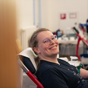Eine Person sitzt auf einer Liege. Ihr wird Blut abgenommen. Sie lacht strahlend in die Kamera