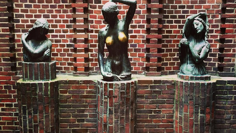 Auf dem Bild sind drei bronzene Frauenbüsten zu sehen, die auf gemauerten Ziegelpfeilern vor einer rotbraunen Backsteinwand stehen. Die mittlere Statue fällt durch ihre glänzend polierten Brüste auf, die auf häufige Berührungen hindeuten. 