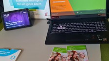 Links ein Tablet, rechts ein Laptop mit einer Präsentation über die Dresdner Seniorenakademie. Vor dem Laptop liegen Flyer. Im Hintergrund steht ein Schild mit der Aufschrift "Wir sind KI-Lernort! Künstliche Intelligenz für ein gutes Altern".