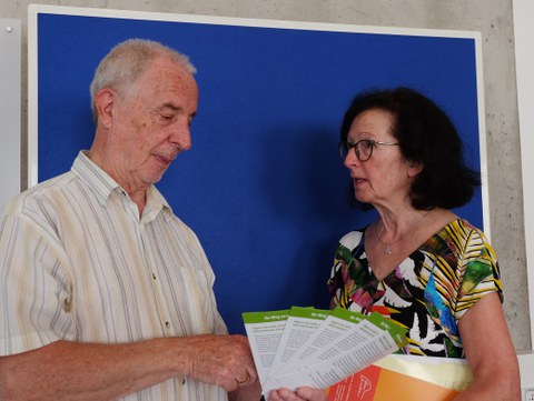 Dieter Seyfarth und Annelie Zeeh stehen vor einer blauen Wand. Annelie Zeeh steht rechtsm spricht und guckt dabei Dieter Seyfarth an.. Dieter Seyfarth steht links und hält Flyer in der Hand, die er betrachtet.