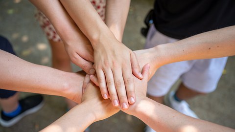 The photo shows a group of students standing in a circle with their hands placed one on top of the other in the center of the circle