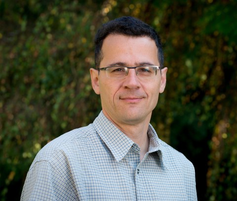 Das Portraitfoto zeigt einen Mann mit kurzem, schwarzem Haar, Brille und blau-kariertem Hemd.