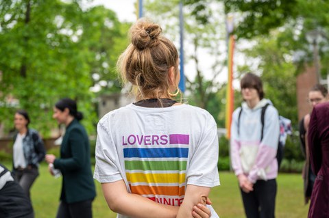 Eine blonde Person ist umgeben von 4 Personen. Man sieht ihren Rücken und sie trägt ein weißes Shirt mit einem bunten Schriftzug, der besagt "Lovers unite"