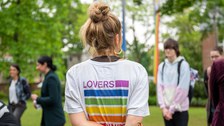 Eine blonde Person ist umgeben von 4 Personen. Man sieht ihren Rücken und sie trägt ein weißes Shirt mit einem bunten Schriftzug, der besagt "Lovers unite"