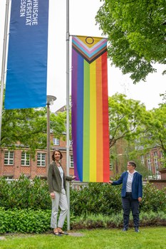 Prorektorin Böhm und Staatssekreträin Dr. Märtens stehen vor der Progressive Pride Flag und halten beide Enden.