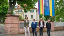 Prorektorin Böhm, Staatssekretärin Märtens, Fay Uhlmann und Carli Wiegand stehen vorm Rektorat, im Hintergrund hängt die Progressive Pride Flag, die TU Dresden Fahne, sowie die Ukrainefahne