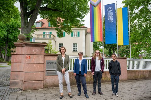 Prorektorin Böhm, Staatssekretärin Märtens, Fay Uhlmann und Carli Wiegand stehen vorm Rektorat, im Hintergrund hängt die Progressive Pride Flag, die TU Dresden Fahne, sowie die Ukrainefahne