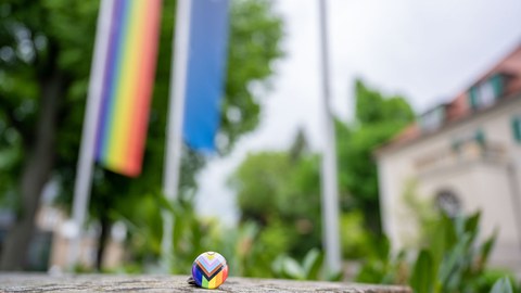 Ein ProgressivePrideFlagButton liegt auf einer Mauer, im Hintergrund ist das Rektorat und die Progressiv Pride Flag zu erkennen