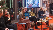 Sechs Frauen sitzen vor einer weihanchtlcihen Kulisse. Die beiden Frauen am Rand gebärdendolmetschen, die Frau in der Mitte hält ein Kinderbuch hoch. 