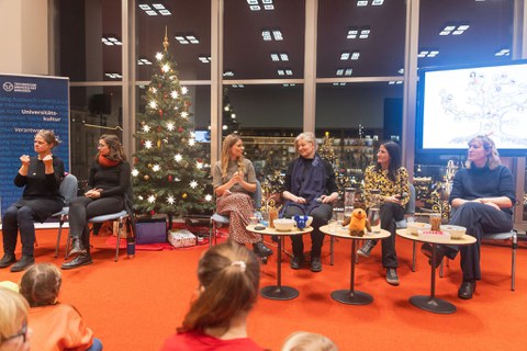 Sechs Frauen sitzen vor einer weihnachtlichen Kulisse auf dem Podium. Die beiden Frauen links gebärdendolmetschen.