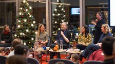 Fünf Frauen sitzen auf dem Podium, eine Person steht hinter IHnen. Links steht ein großer Weihnachtsbaum. 
