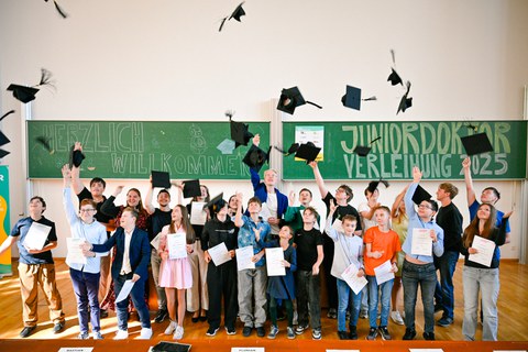 Ca. 25 Kinder und Jugendliche stehen in 2 Reihen vor einer grünen Tafel und werfen Doktorhüte hoch. Auf der Tafel steht "Herzlich willkommen. Juniordoktor-Verleihung 2025."