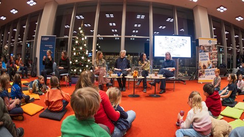 Ein Podium mit 4 Personen, davor sitzen Kinder auf dem Boden. Neben dem Podium 2 Gebärdensprachdolmetscherinnen, hinter ihnen ein beleuchteter Nadelbaum.