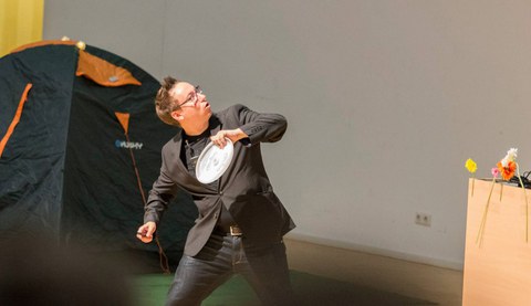 Das Bild zeigt eine männliche Person in Jeans, T-Shirt und Jacket. In der linken Hand hält er ein Frisbee und ist dabei, es zu werfen. 