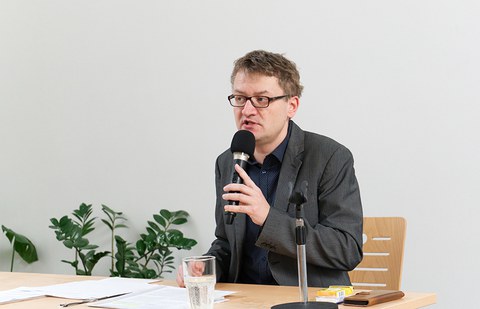 Das Foto zeigt einen Mann mit Mikro in der rechten Hand. Er sitzt an einem Tisch. Vor ihm auf dem Tisch ein Glas Wasser und A4-Blätter.