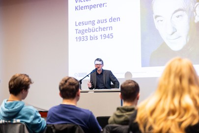A man sitting on a stage and speaking to people sitting in front of him.