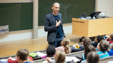 A man speaks to children in a lecture theatre. The children sit on lecture theatre chairs with a writing tablet and look at him.