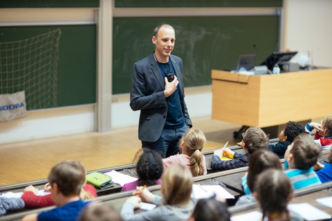A man speaks to children in a lecture theatre. The children sit on lecture theatre chairs with a writing tablet and look at him.
