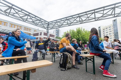 The photo shows people sitting on benches in a marquee. The benches are arranged in several rows. One of the people sitting there raises their hand. Another person in the audience gestures to draw attention to the person who has raised their hand.